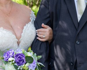 Bride in lace gown holding purple rose bouquet, linked arm with groom in gray suit, showing diamond engagement ring.
