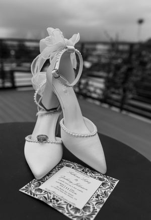 Elegant white pearl-strapped bridal heels with floral bow displayed beside wedding invitation and ring in black and white photo.
