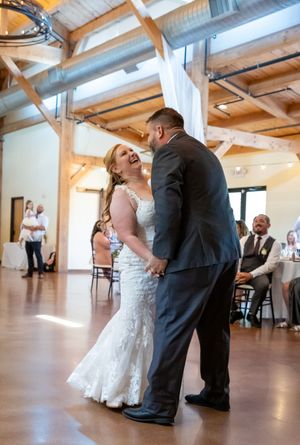 Bride and groom share their first dance, laughing joyfully in a rustic wedding venue with wood beam ceilings.