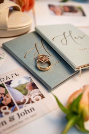 Wedding rings rest on sage green vow books beside a save the date card for John and his bride, with bridal heels nearby.