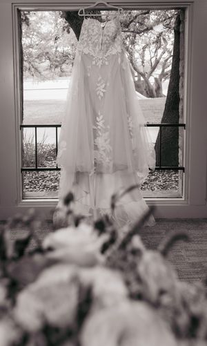 Black and white photo of a lace wedding gown hanging in a window with floral bouquet in foreground.