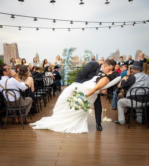 Groom dips bride for a romantic kiss at an outdoor rooftop wedding ceremony with city skyline views.