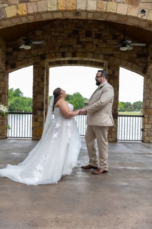 Bride and groom holding hands under a stone arch pavilion overlooking a lake on their wedding day.