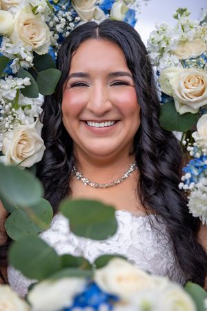 Smiling bride with dark curly hair wearing a crystal necklace, surrounded by white roses and blue flowers.