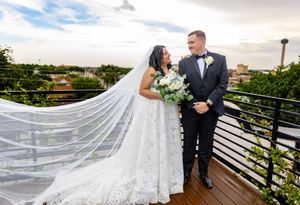 Bride and groom share a romantic moment on a rooftop terrace, her lace gown and veil flowing in the breeze.