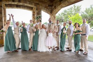 Wedding party in green and tan attire celebrating under stone archway with bride and groom at center.