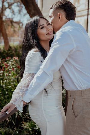 A romantic couple embraces outdoors, with the woman wearing a white lace dress against a garden backdrop.