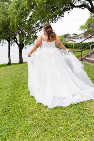Bride in white lace ball gown holding her train on green grass near a lake, viewed from behind.
