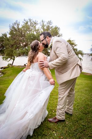 Bride and groom share a romantic kiss outdoors on green grass near a waterfront, bride in white ballgown, groom in tan suit.