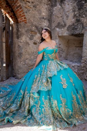 Young woman in a teal and gold quinceañera ball gown with tiara poses in a rustic stone archway.
