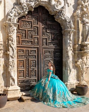 Young woman in teal gold quinceañera ball gown poses before ornate carved wooden doors of historic stone building.