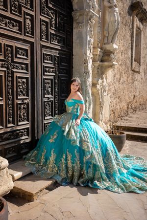 Young woman in a stunning turquoise and gold quinceañera ball gown posing by ornate wooden doors.