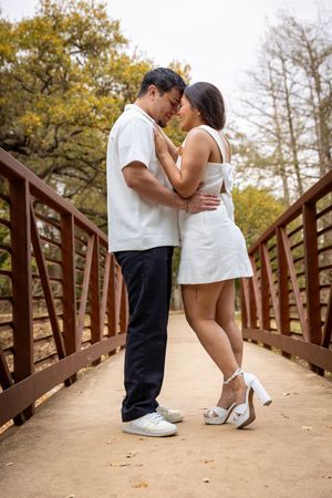 Couple in white outfits embracing on a rustic red bridge surrounded by autumn trees.