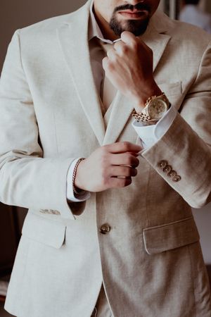 Man in beige linen suit adjusting cufflinks wearing gold watch and bracelet, elegant formal attire.