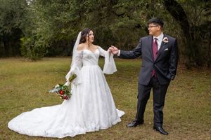 A couple in wedding attire stands together in a wooded outdoor setting during their special day.