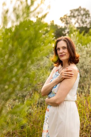 Woman in floral white dress standing outdoors among green bushes, smiling gracefully at camera.