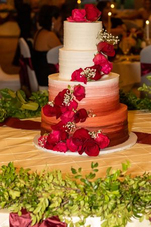 A three-tiered wedding cake decorated with deep red roses and baby's breath sits on a wooden table with greenery.