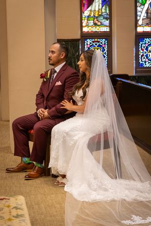 A bride and groom sit together in a church with stained glass windows while wearing burgundy and white wedding attire.