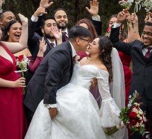 Bride and groom share a kiss surrounded by cheering wedding party in burgundy and black attire outdoors.