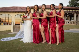 A wedding party in burgundy dresses poses together on a grassy lawn near a Spanish-style building.