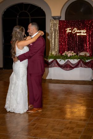A couple shares a romantic first dance on a wooden dance floor with red and gold decor in the background.