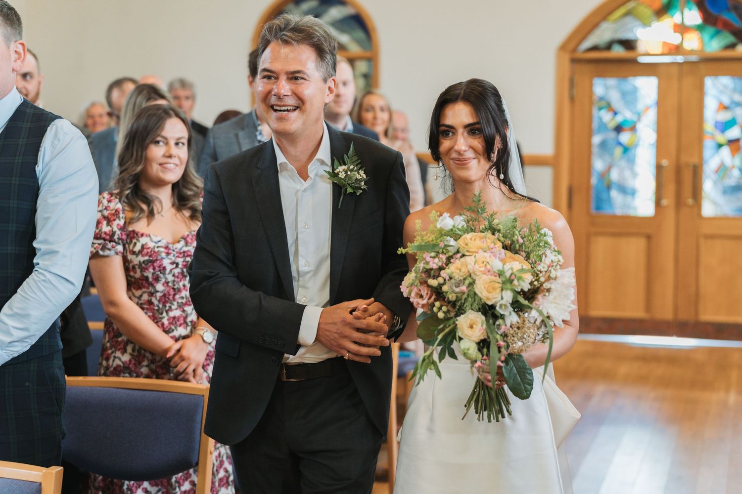 Wedding guests mingle and socialize during a reception in a bright wooden interior venue with floral arrangements.