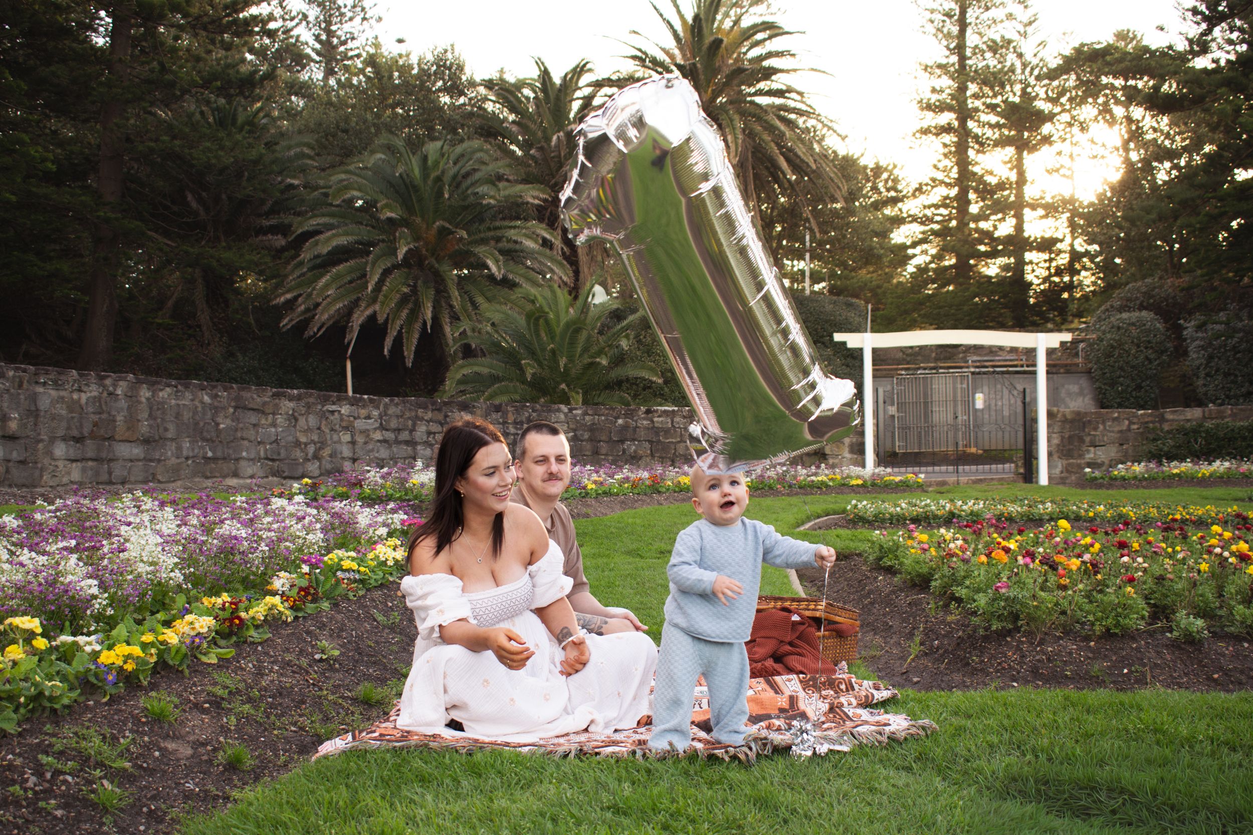 Mum and dad with their toddler, number 1 helium balloon, golden hour in a Newcastle flower garden during 1st birthday milestone photoshoot