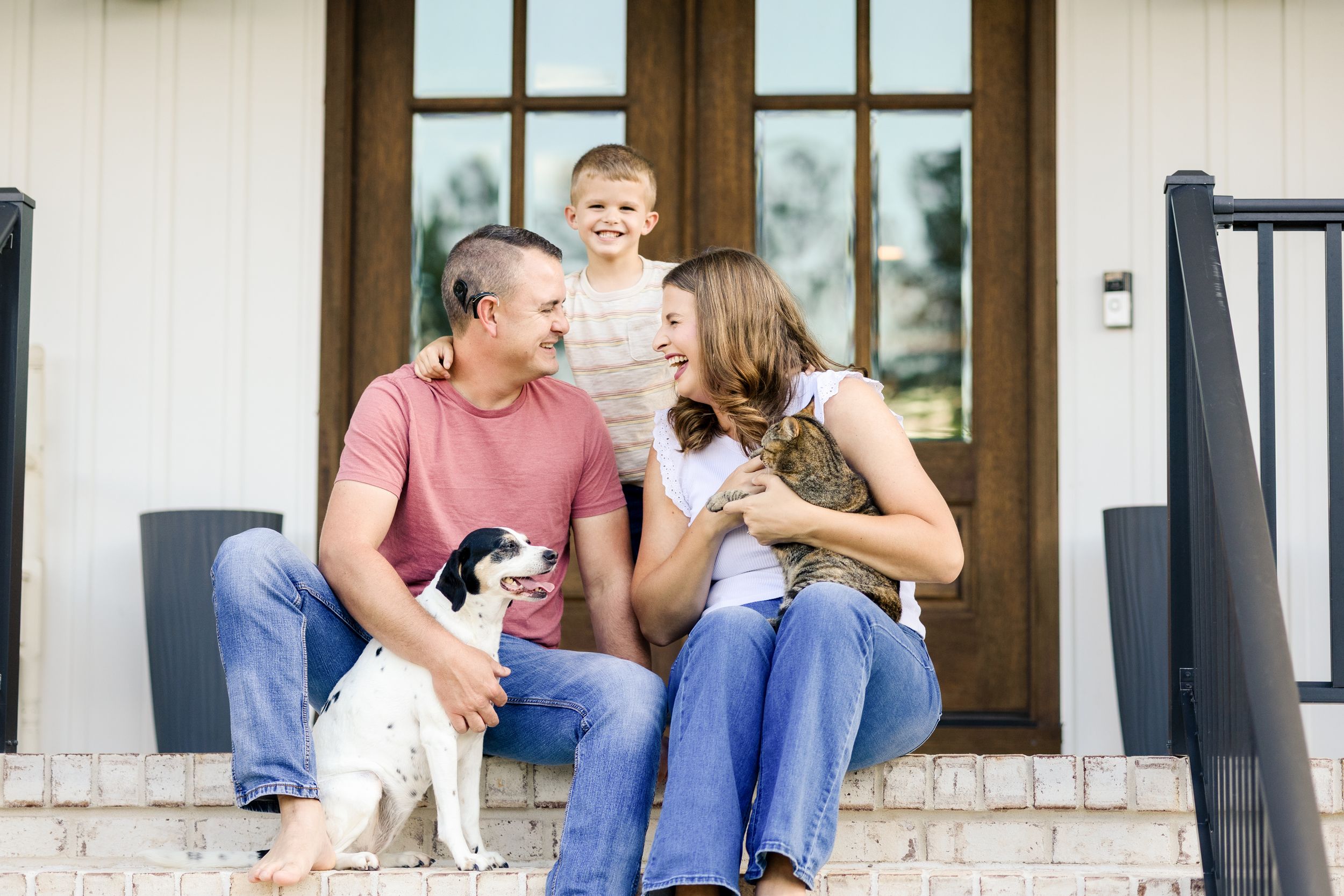 A couple sits together on a porch step with their small white dog, sharing joyful moments outdoors.