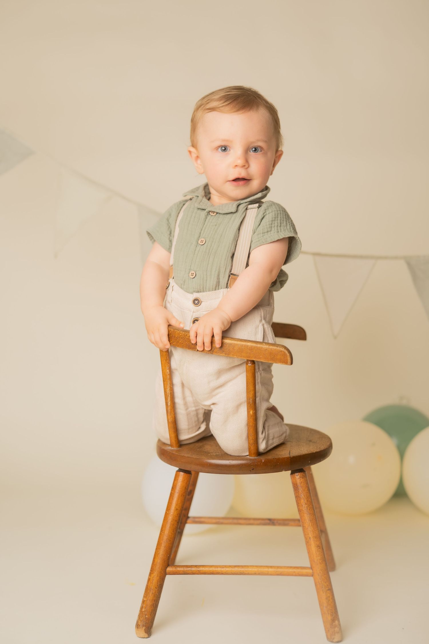 Small child in sage green shirt and beige pants stands on wooden chair against neutral backdrop with party decorations.