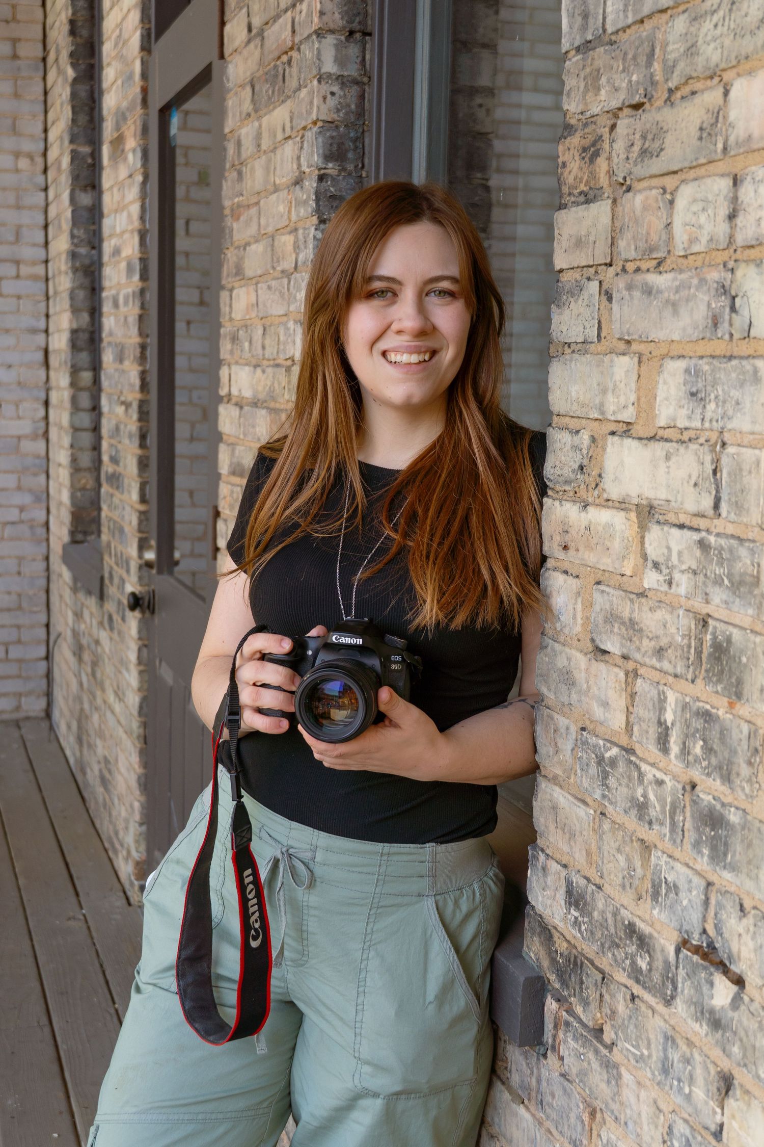 A photographer in a gray sweater and jeans holds up a camera while posing outdoors in an autumn setting.