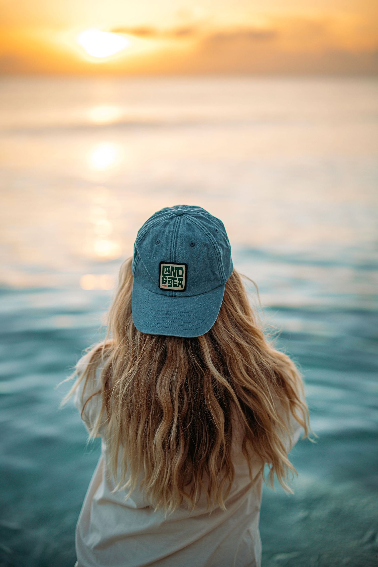 Person wearing blue cap looking out at sunset over ocean waves.