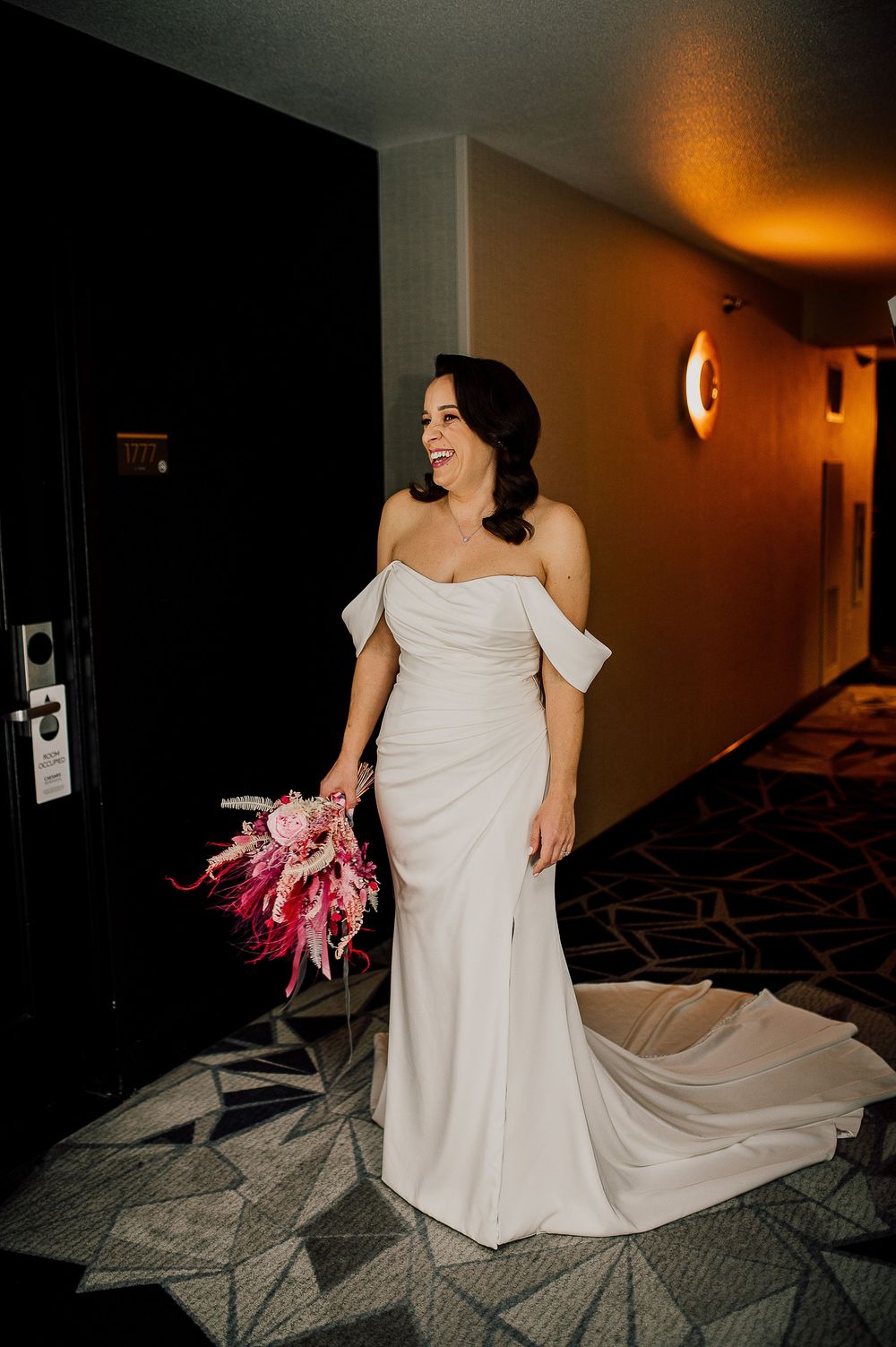 A person in a flowing white wedding dress twirls playfully in a dimly lit hotel hallway with carpeted floors.