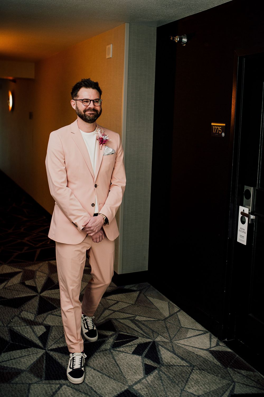 A groom in a light pink suit poses in various angles along a hotel corridor with geometric patterned carpet.