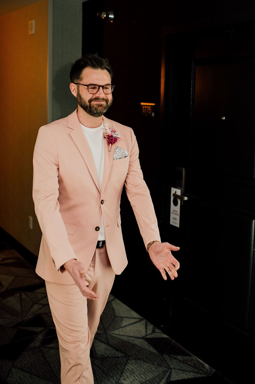 A person wearing a light pink suit and glasses gestures while standing in a dimly lit hallway.