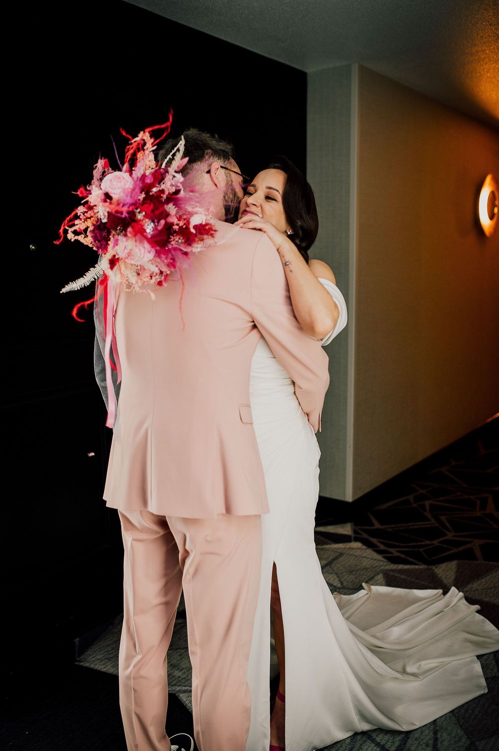 Two wedding attendees embrace in a tender moment, one wearing a white gown and the other in a pink suit.