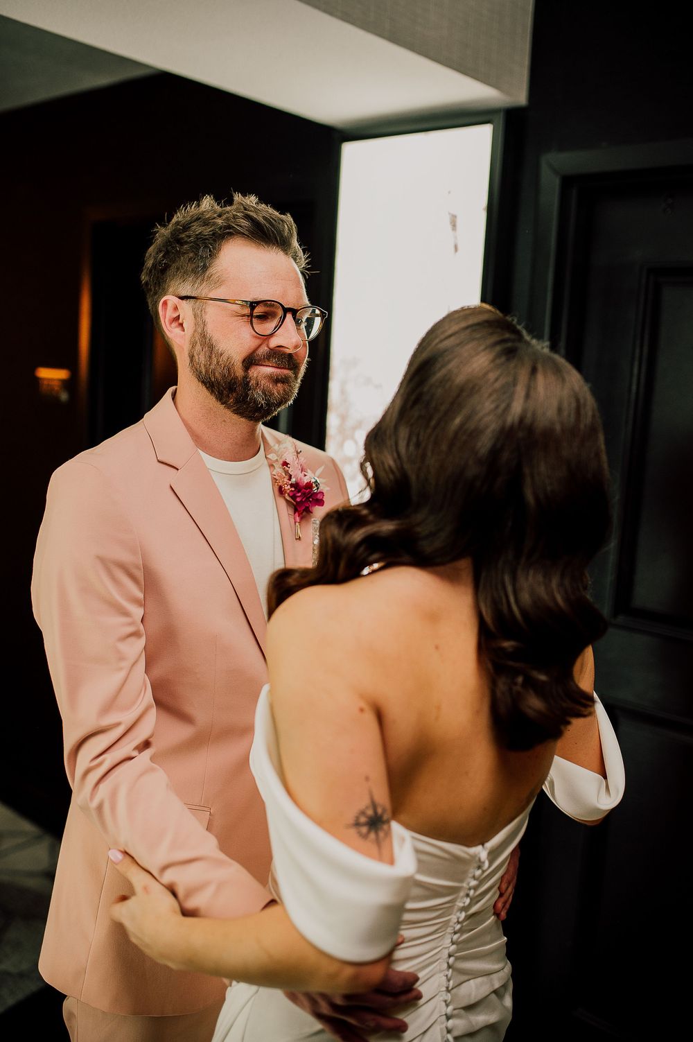 A couple shares an intimate moment at their wedding, with the groom wearing a pink suit and the bride in a white dress.