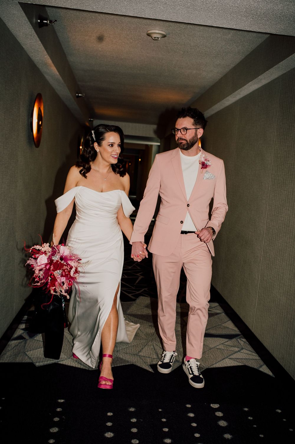 A couple in wedding attire walks through a dark hallway, with the bride in white and groom in pink suit.