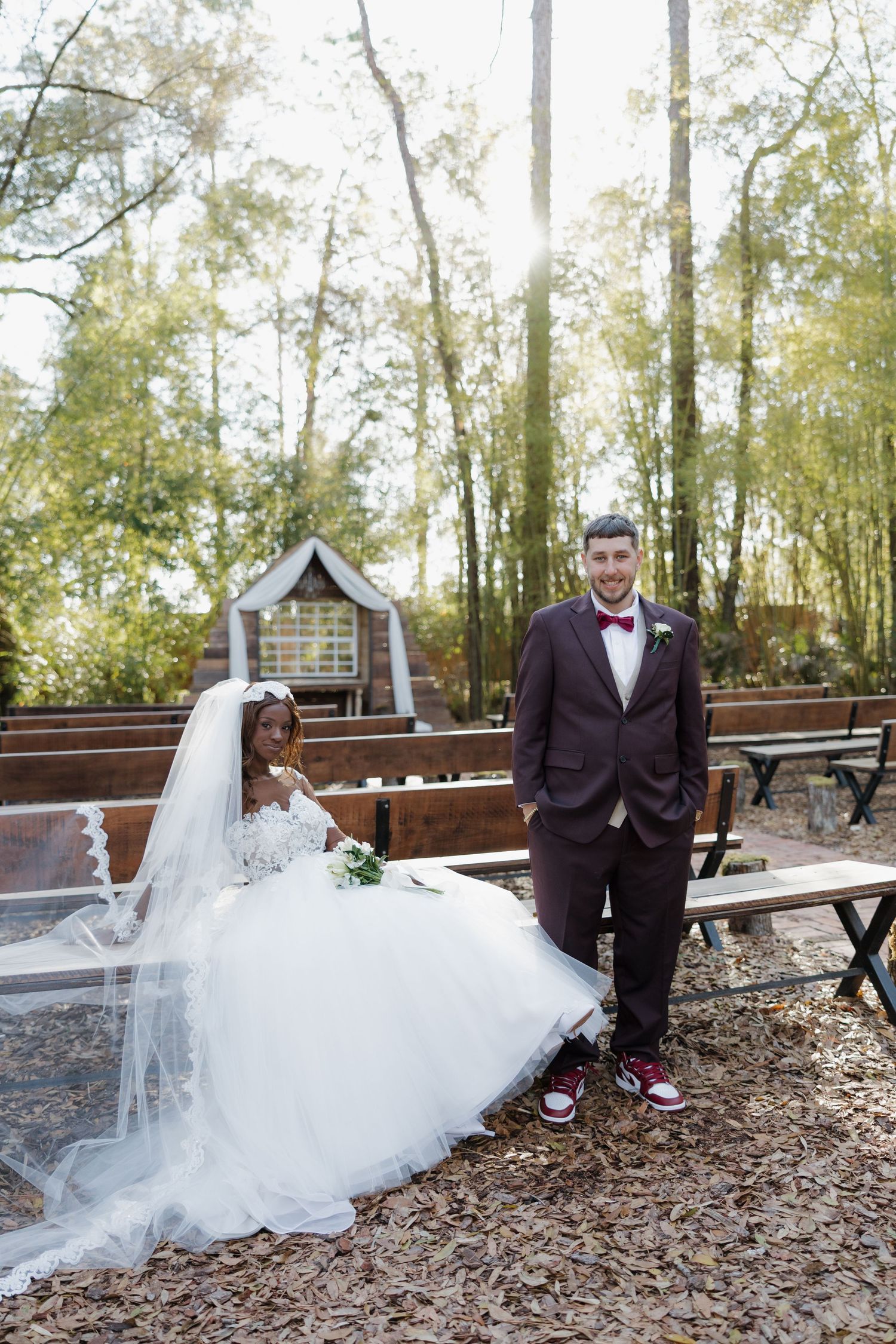 Bride in flowing white dress and groom in tuxedo pose on wooden walkway surrounded by trees.