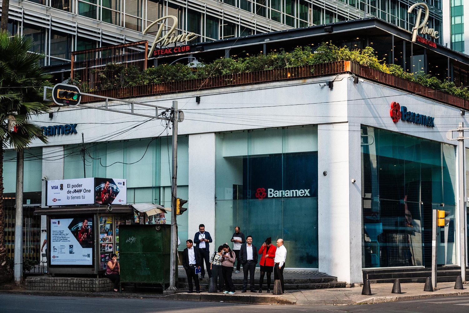 People wait in line outside a bank branch with glass windows and signage on a city street.