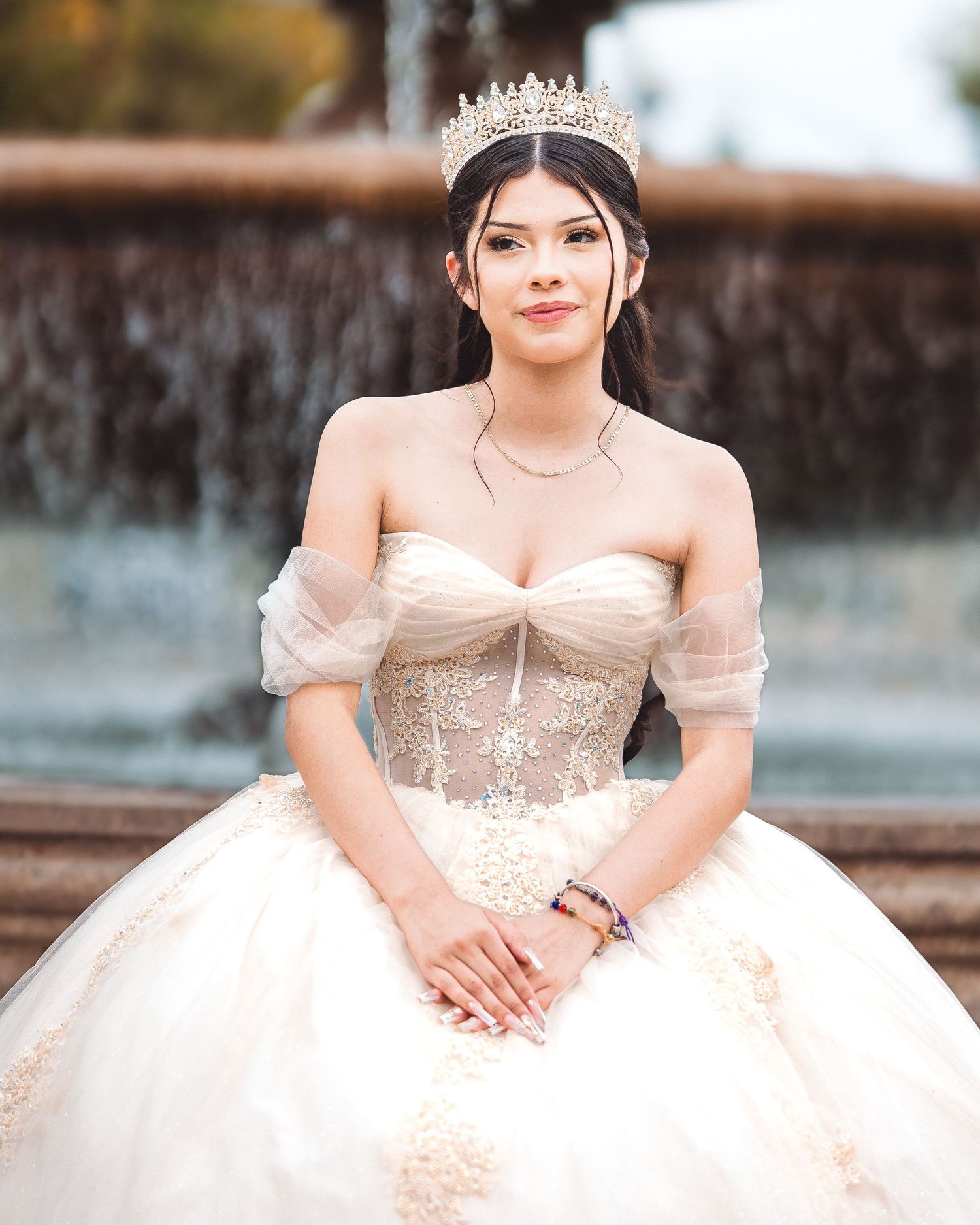 A bride in an elegant white ballgown poses by a cascading waterfall fountain in an outdoor setting.