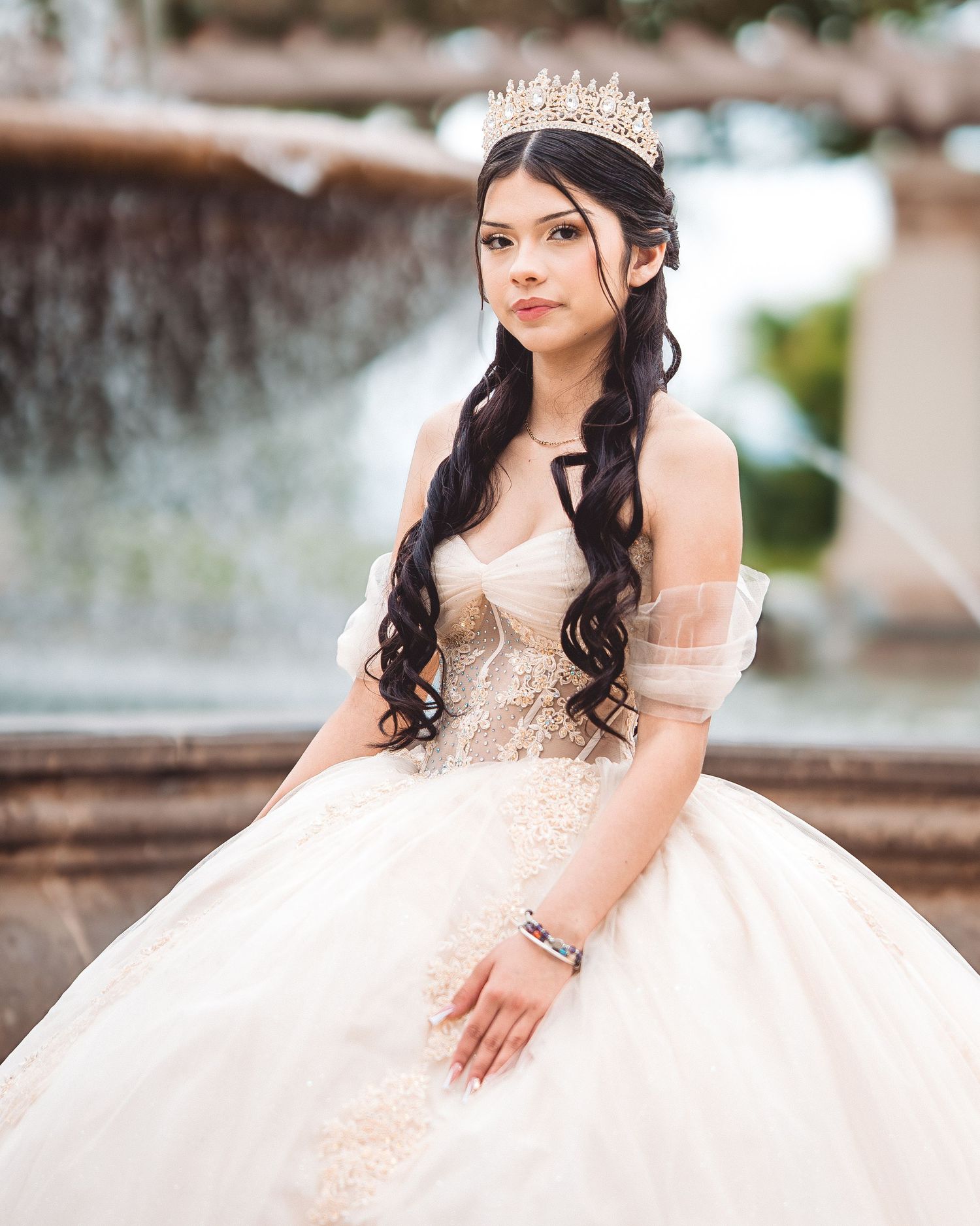 A person in an elegant champagne-colored quinceañera ball gown with a tiara poses in front of a fountain.