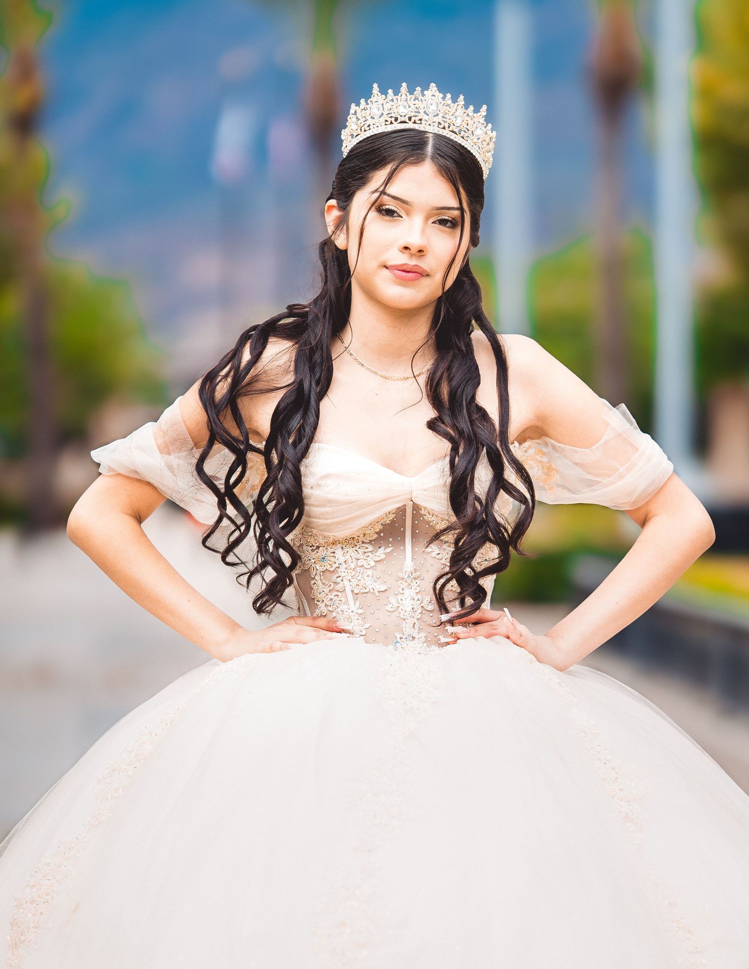 Young woman in a white princess ball gown and tiara poses gracefully in an outdoor setting with blurred greenery background.