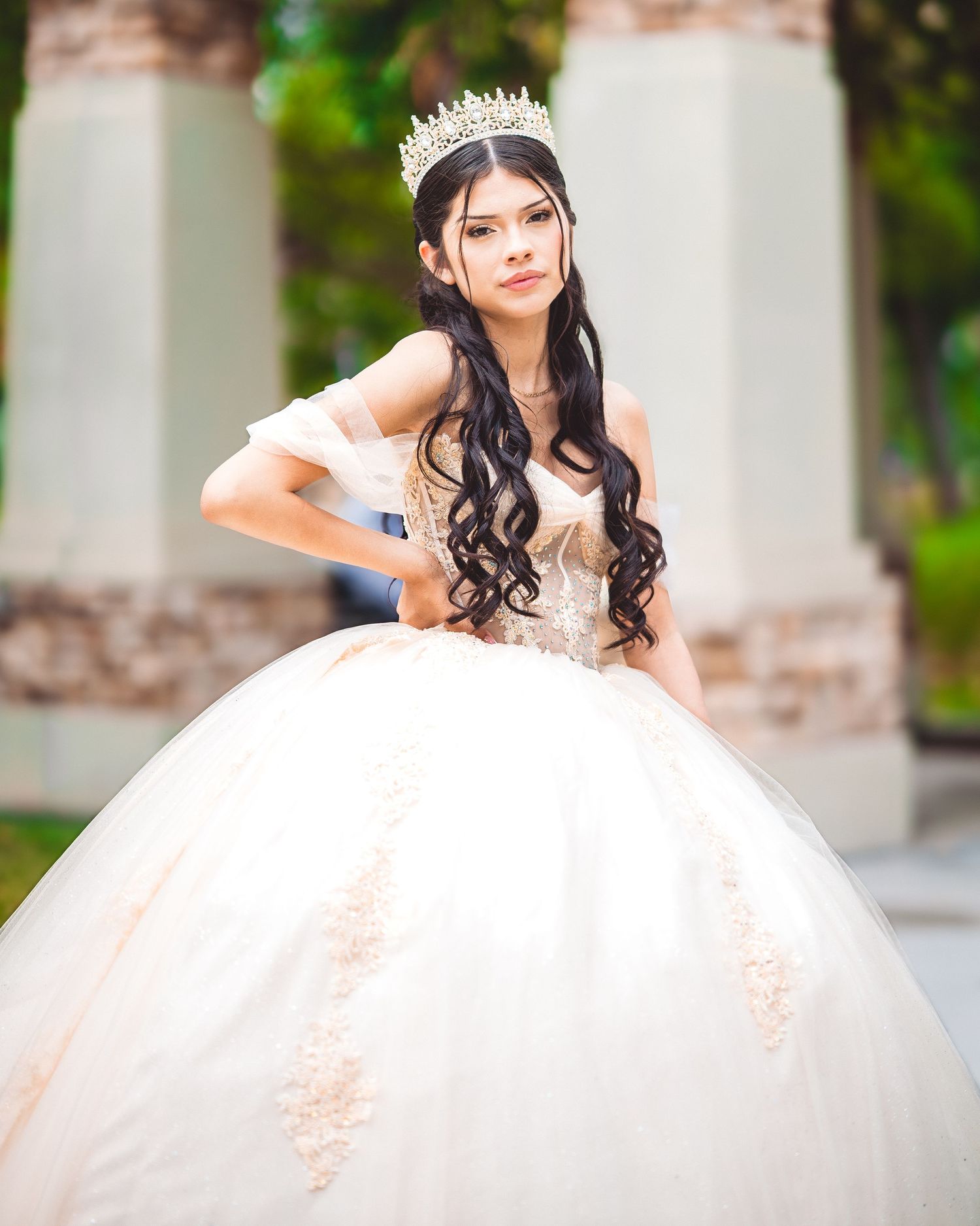 A bride in a white ballgown poses gracefully in an elegant outdoor setting with stone columns and greenery.