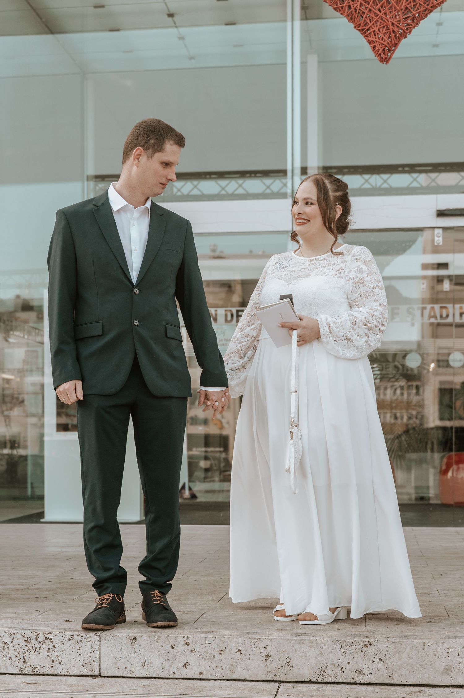 A bride in a white lace gown and groom in a dark green suit hold hands and smile at each other outdoors.