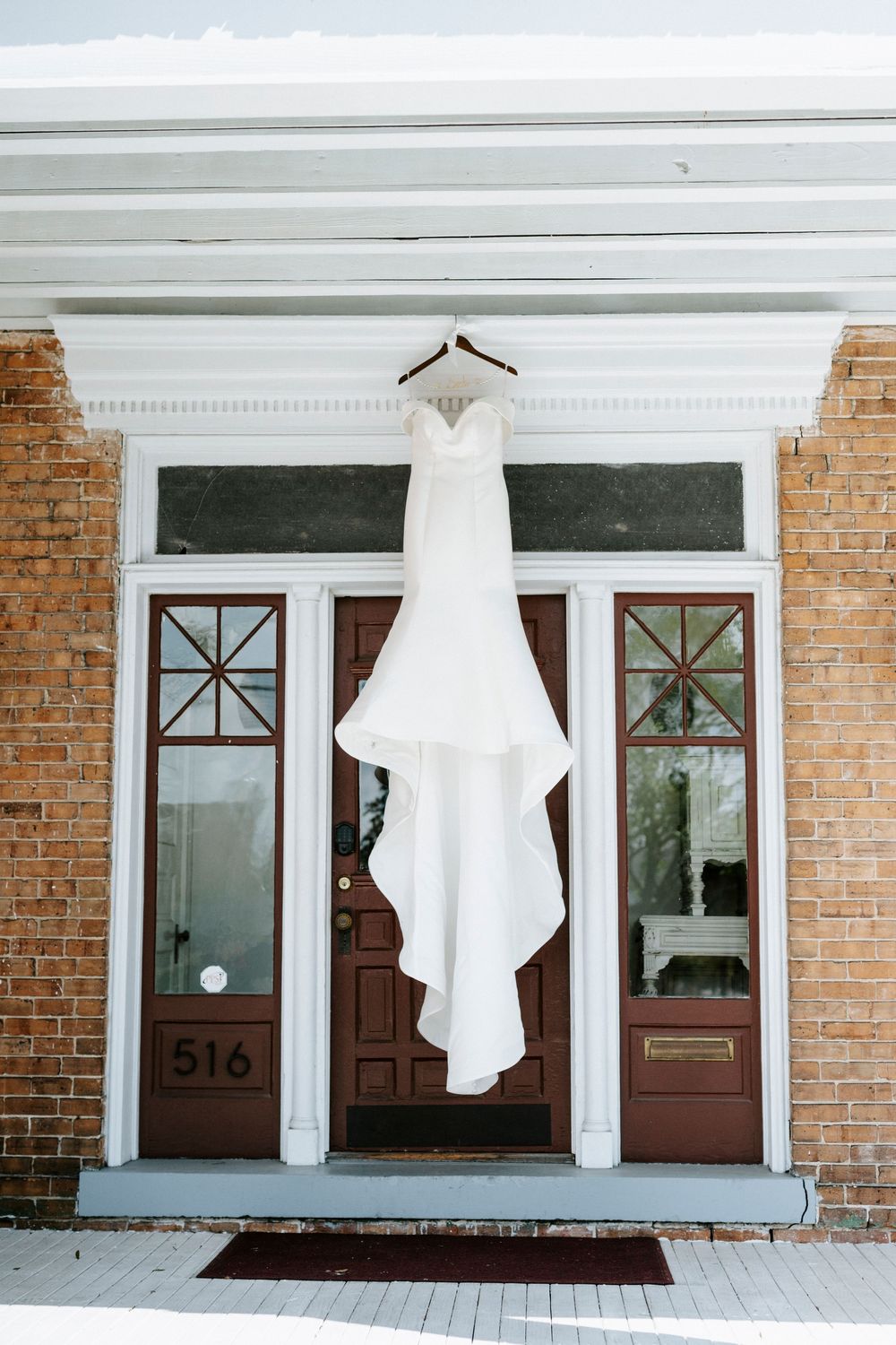 White wedding dress hangs elegantly against brick exterior with brown doors and white columns at venue 516.