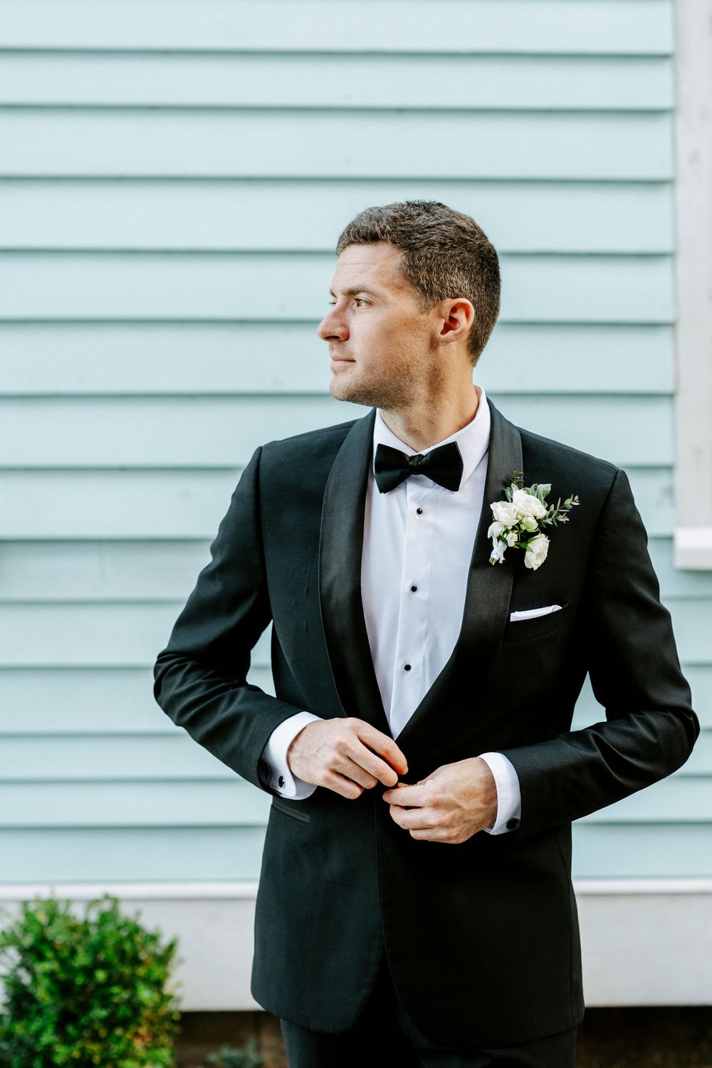A series of elegant black tuxedo portraits against a white siding backdrop with a small boutonniere.