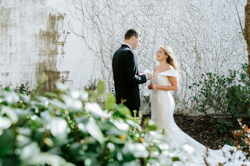 Wedding couple poses against white textured wall with lush greenery.