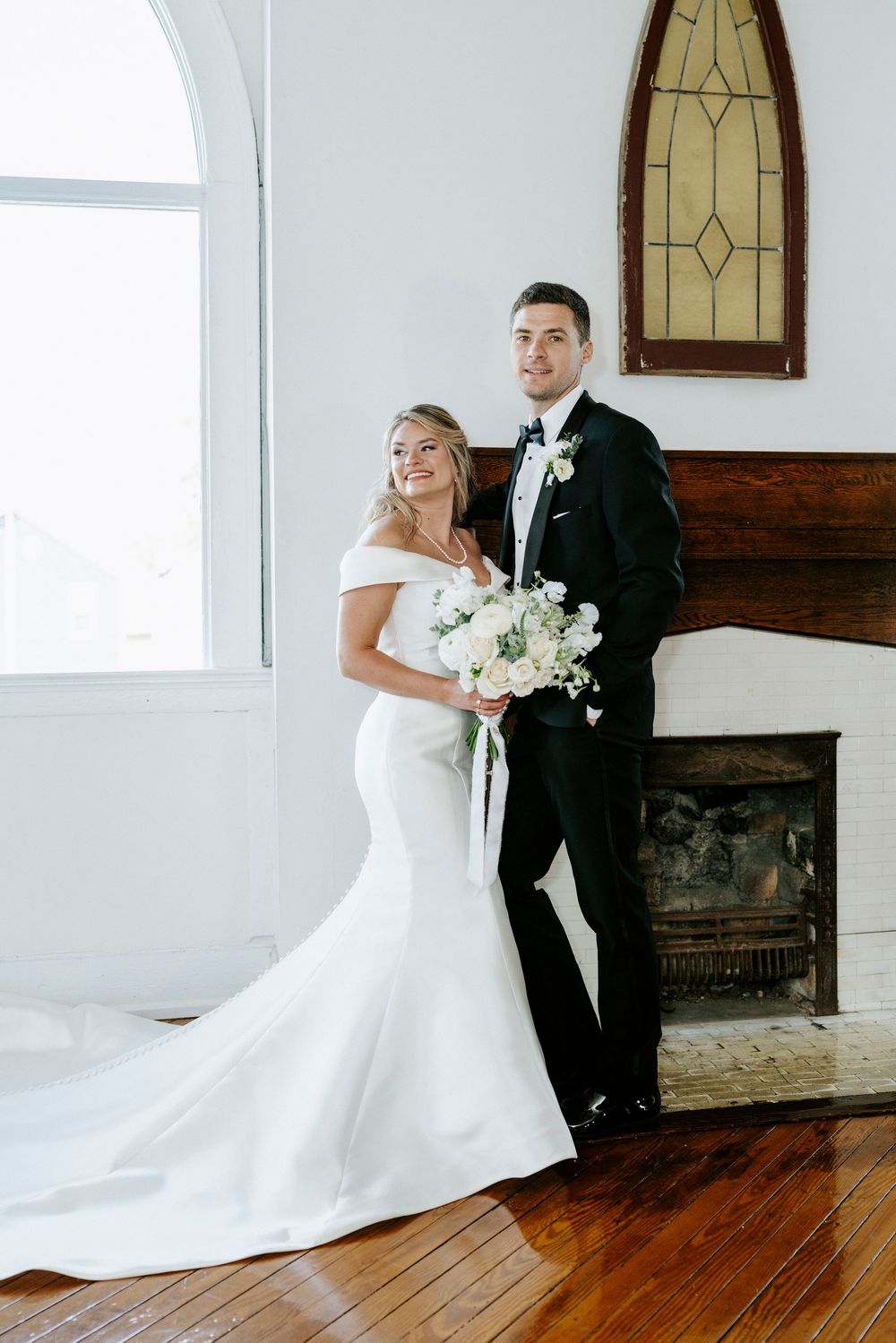A bride in a white off-shoulder mermaid gown and groom in black tuxedo pose in front of a rustic fireplace.