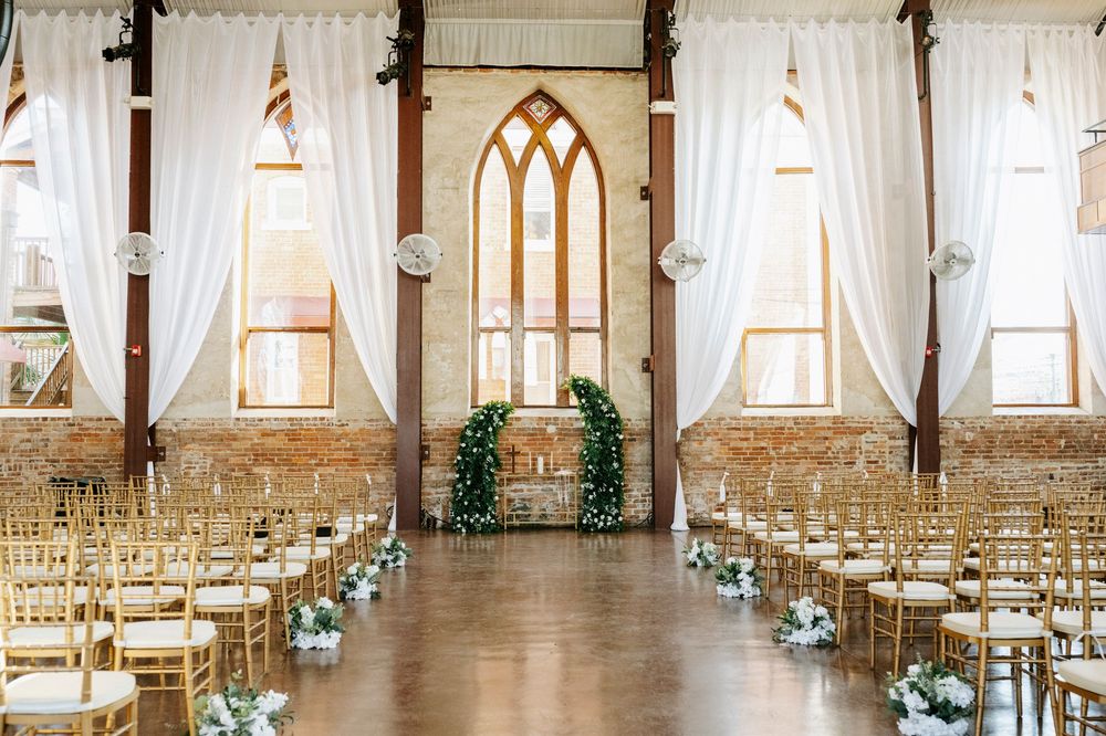 Elegant wedding venue interior with cathedral windows, white drapes, and wooden chairs set for ceremony.
