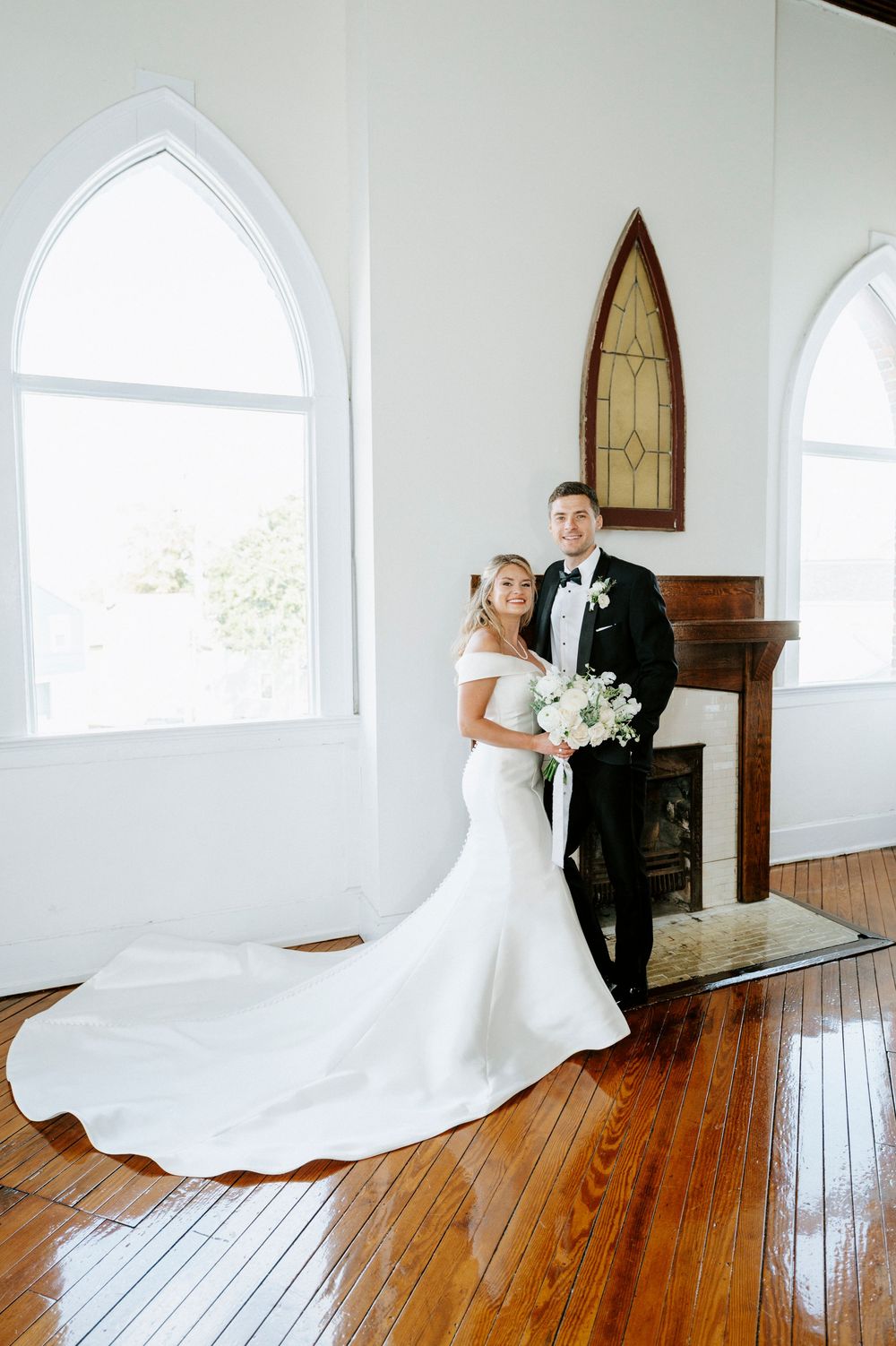 Wedding couple poses in chapel with arched windows and hardwood floors.